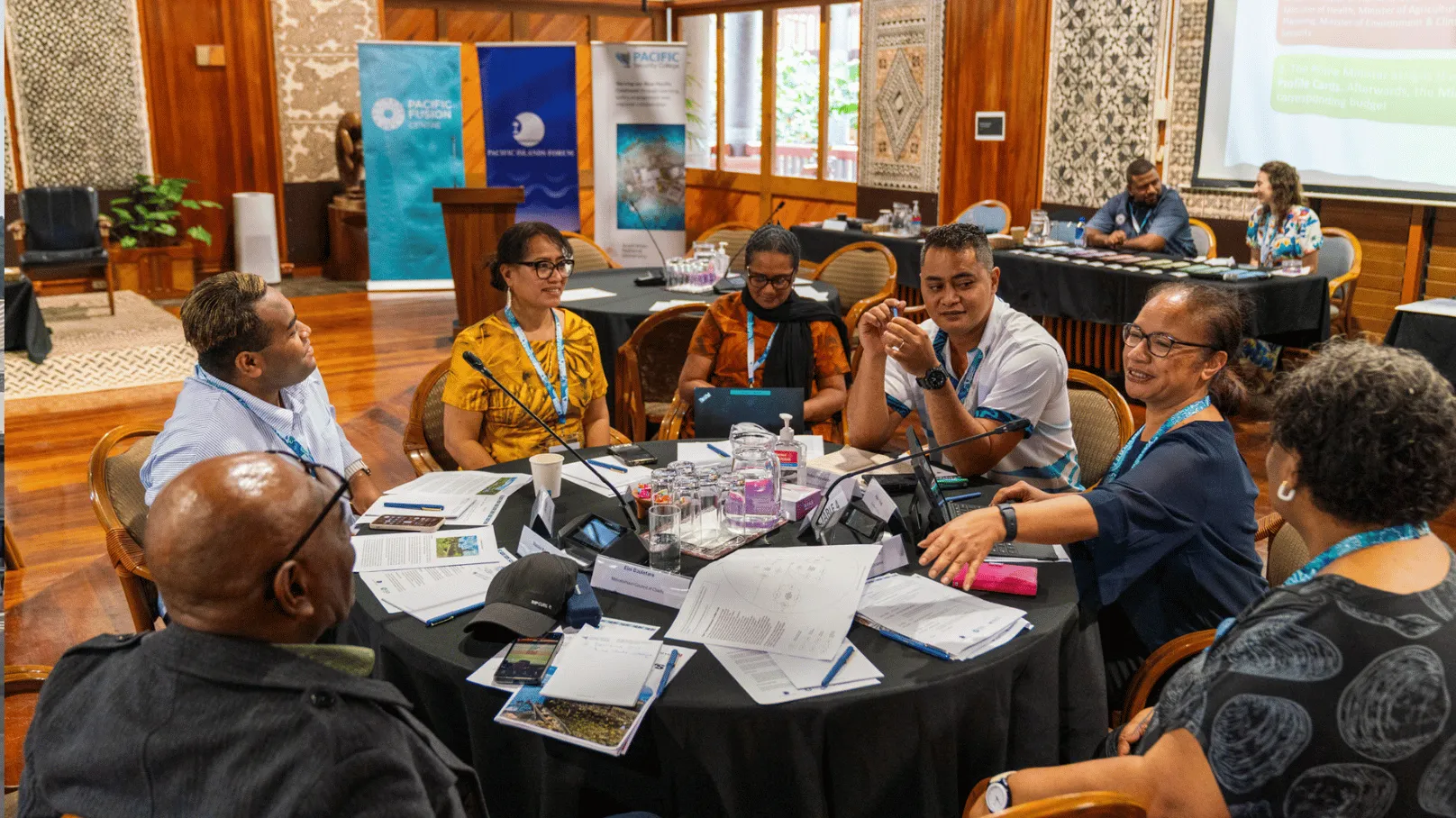 Participants sit around a round table during a workshop session, engaged in discussion and collaborative problem-solving. Documents, laptops, notebooks and refreshments are spread across the table, while presentation screens and event banners are visible in the background.