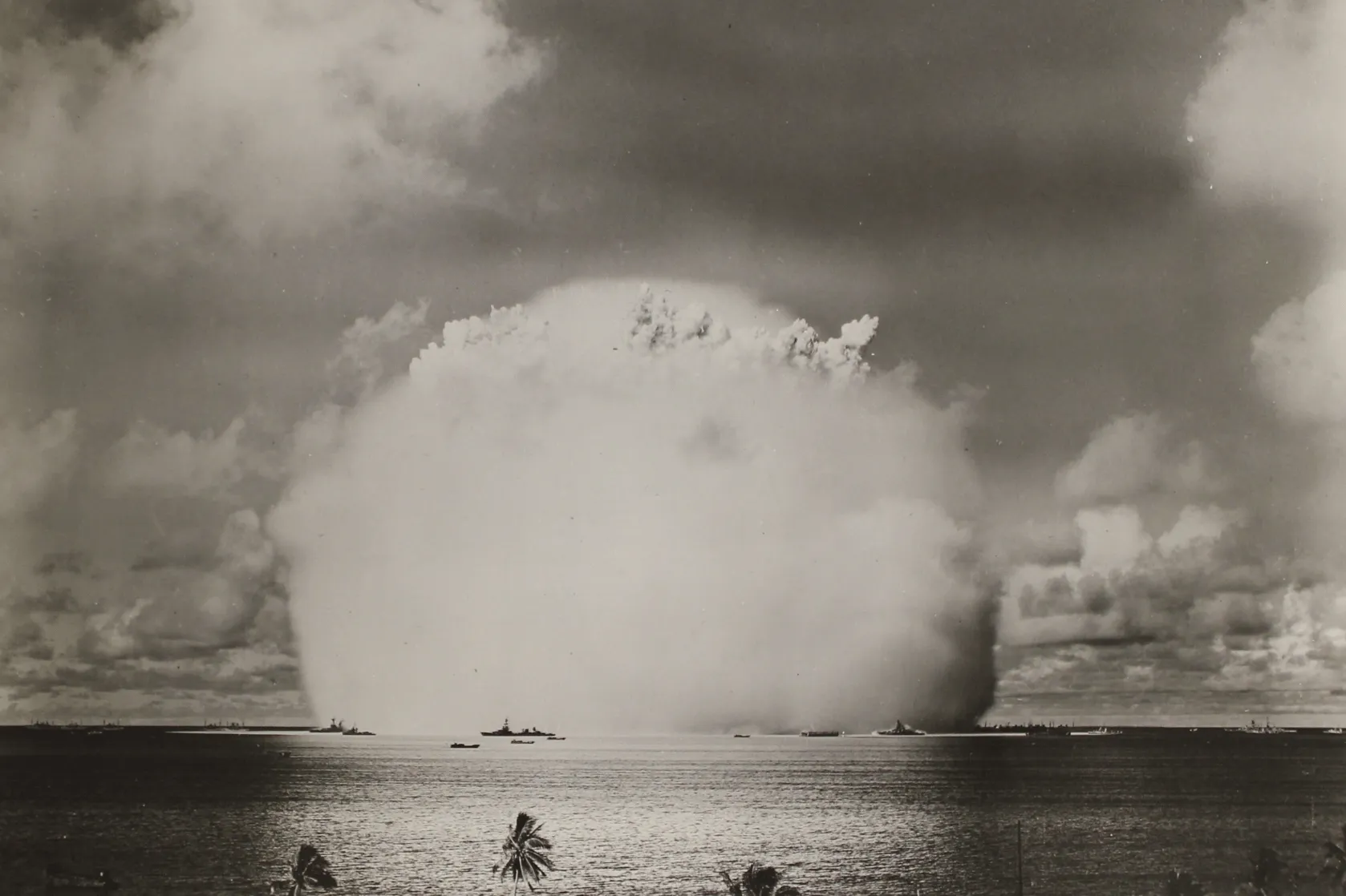 Black-and-white photograph of a massive mushroom cloud rising from an atmospheric nuclear test over the ocean. A towering column of water and spray dominates the centre of the image, with several naval ships positioned around the blast at a distance. Dark clouds fill the sky above, while a few palm trees are visible in the foreground, emphasising the immense scale of the explosion.