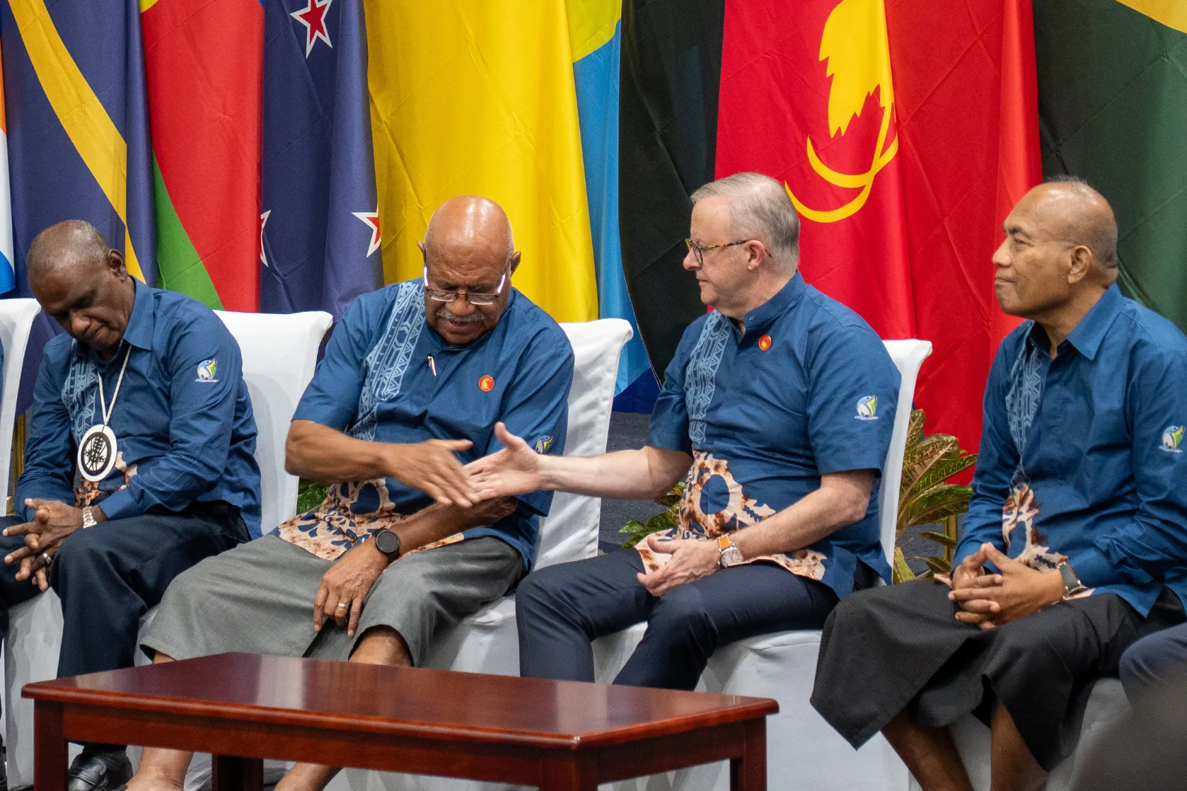 Four leaders wearing matching blue Pacific-themed shirts sit on a stage in front of national flags during a regional event. Two of the leaders are shaking hands while the others look on, with a coffee table positioned in front of them, conveying a moment of cooperation and dialogue.