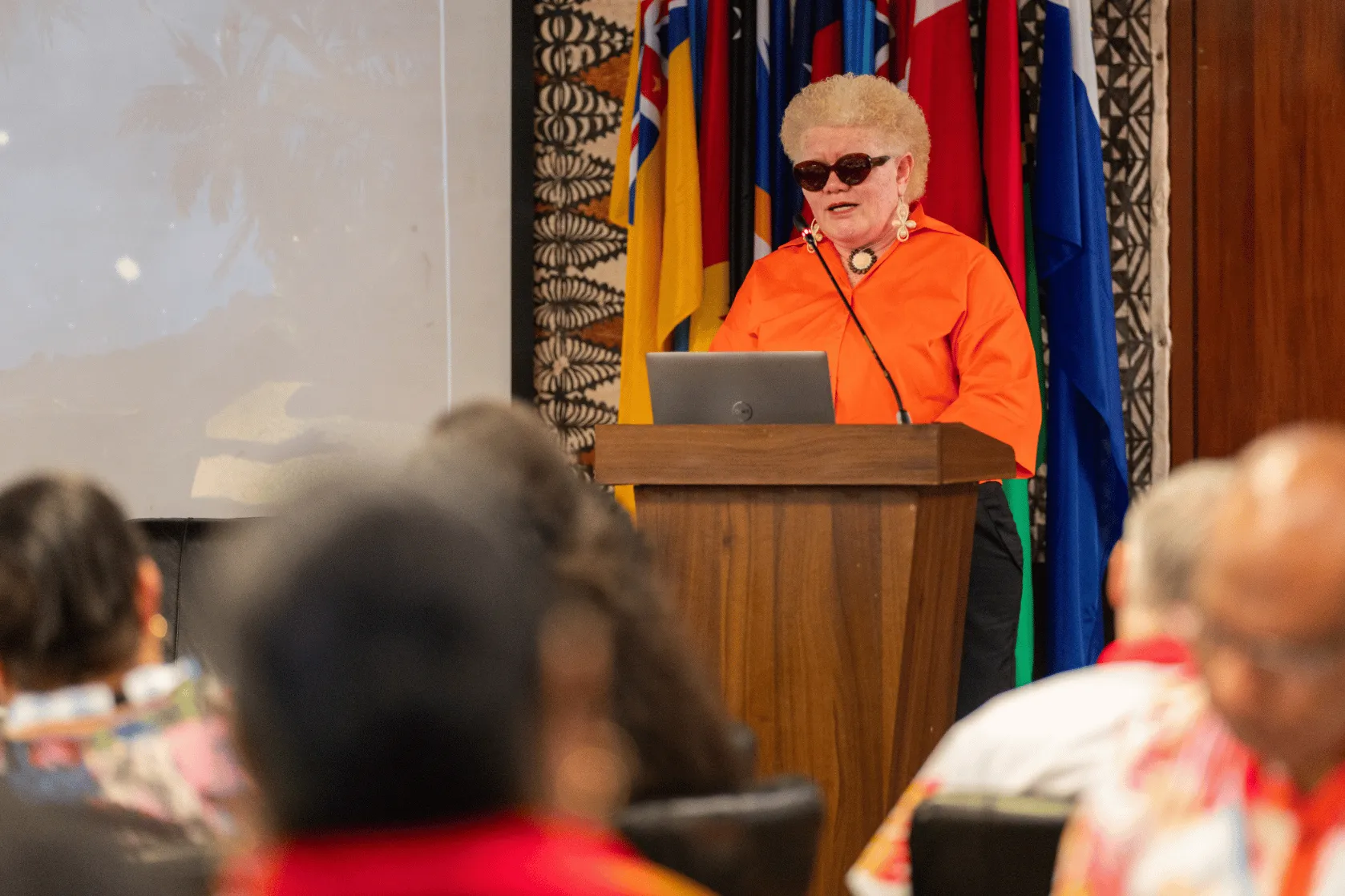 A person wearing a bright orange shirt and dark sunglasses stands at a lectern delivering a presentation, with a laptop and microphone in front of them. A row of Pacific national flags forms the backdrop while audience members are visible out of focus in the foreground.