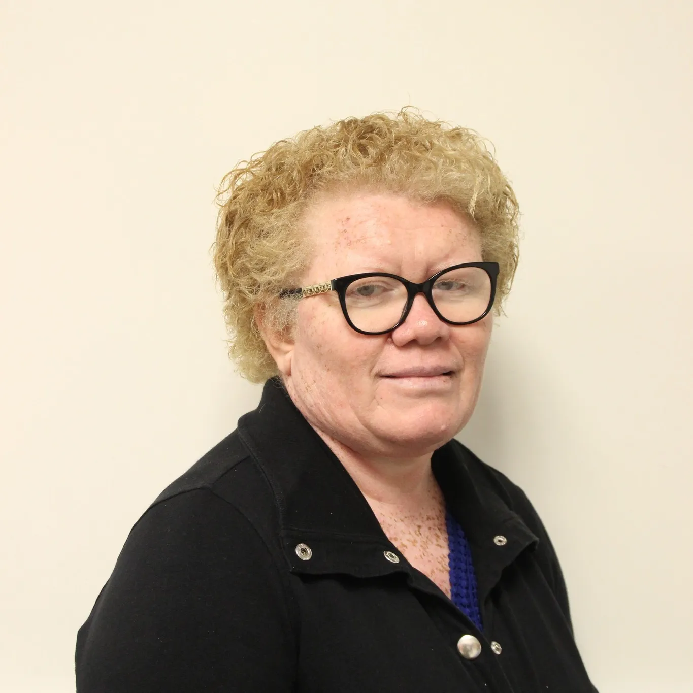 Portrait of a person with short, curly blond hair and black-framed glasses, wearing a black jacket over a blue top. They are facing slightly to the side and smiling gently against a plain cream-coloured background.