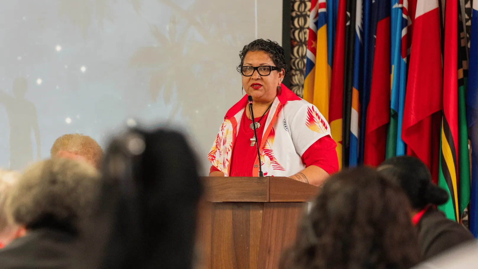 A speaker stands at a lectern addressing an audience at a conference, with Pacific national flags displayed behind them. The speaker wears a red and white Pacific-inspired shirt and speaks into a microphone while attendees are seated in the foreground.