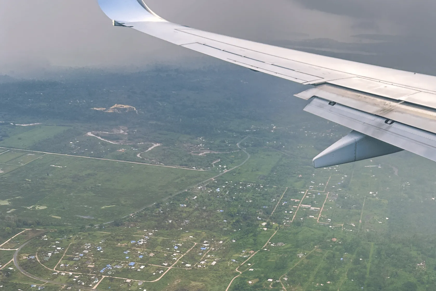 View from an airplane window showing the aircraft wing above a lush green landscape dotted with homes, roads and winding waterways. Low, dark clouds hang over the horizon, with rain visible in the distance as the plane descends.