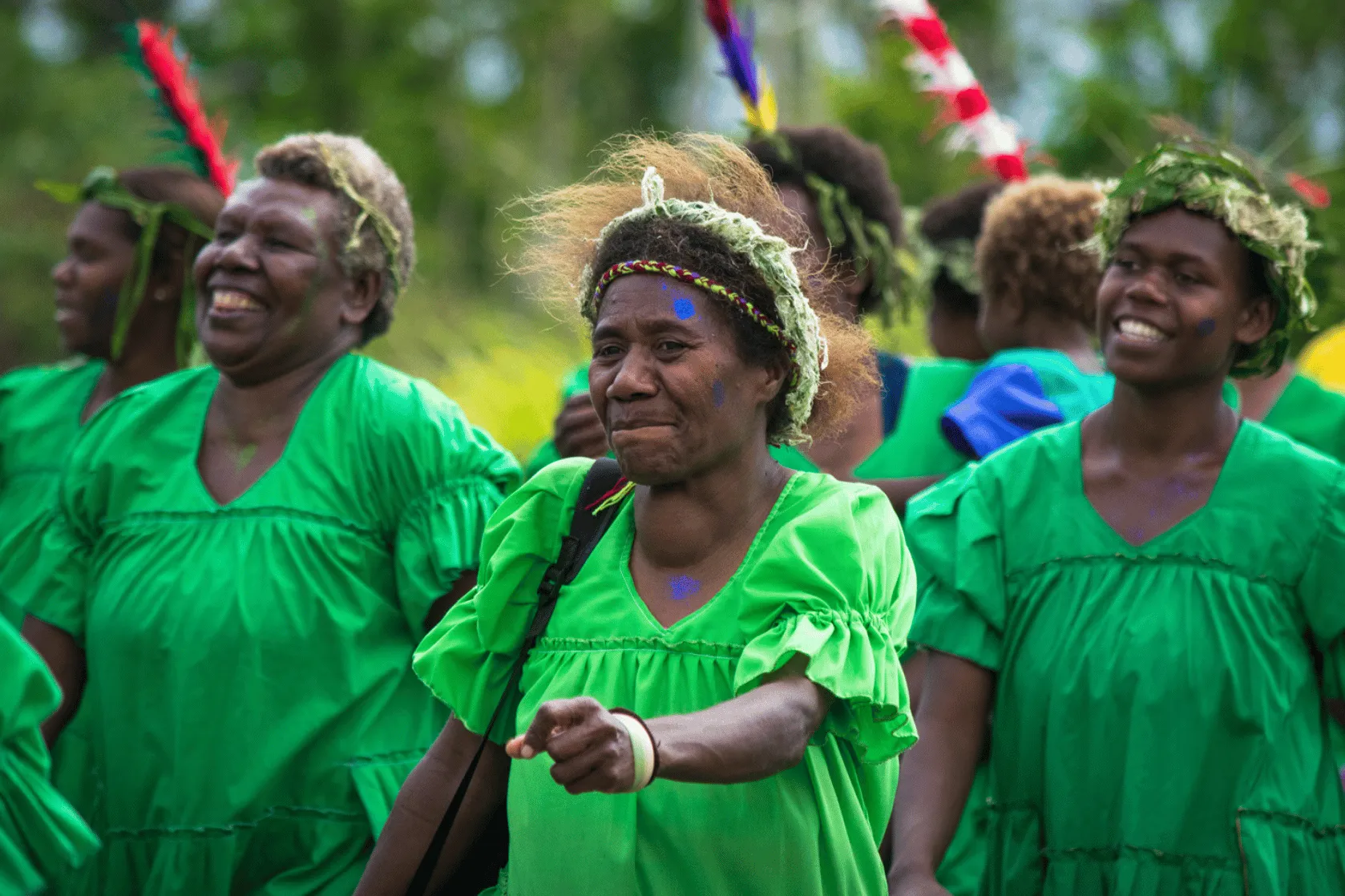 A group of women in bright green dresses smile and dance during a cultural celebration outdoors. Several wear woven leaf headpieces, face paint and colourful feathered headdresses, with lush greenery blurred in the background.