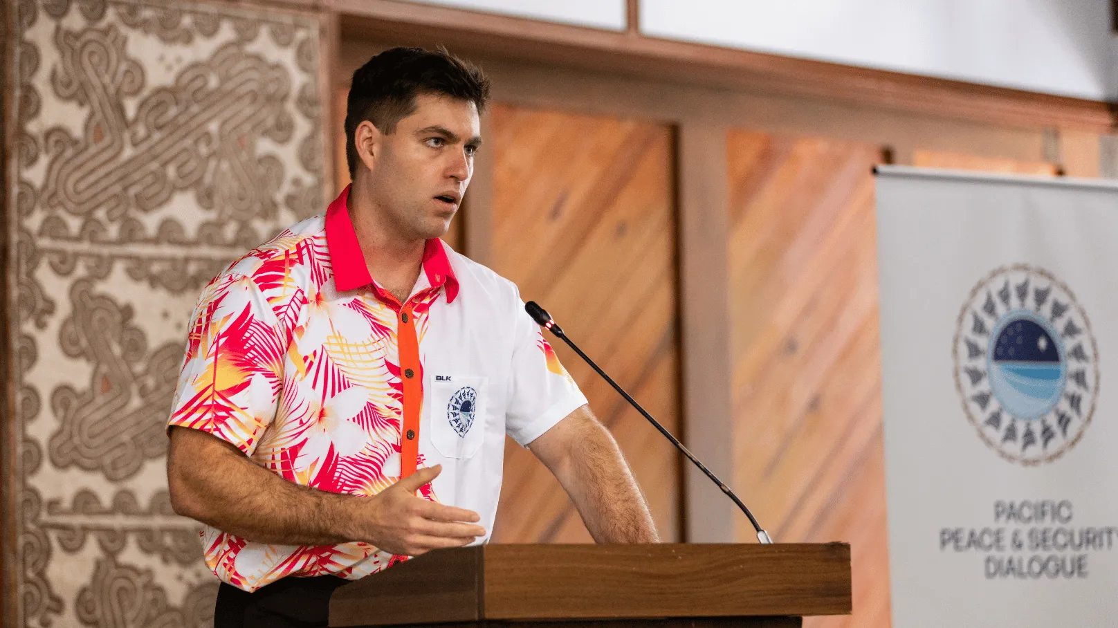 A speaker stands at a lectern addressing an audience at the Pacific Peace & Security Dialogue. The speaker wears a bright floral Pacific-style shirt and speaks into a microphone, with a Pacific Peace & Security Dialogue banner visible in the background.