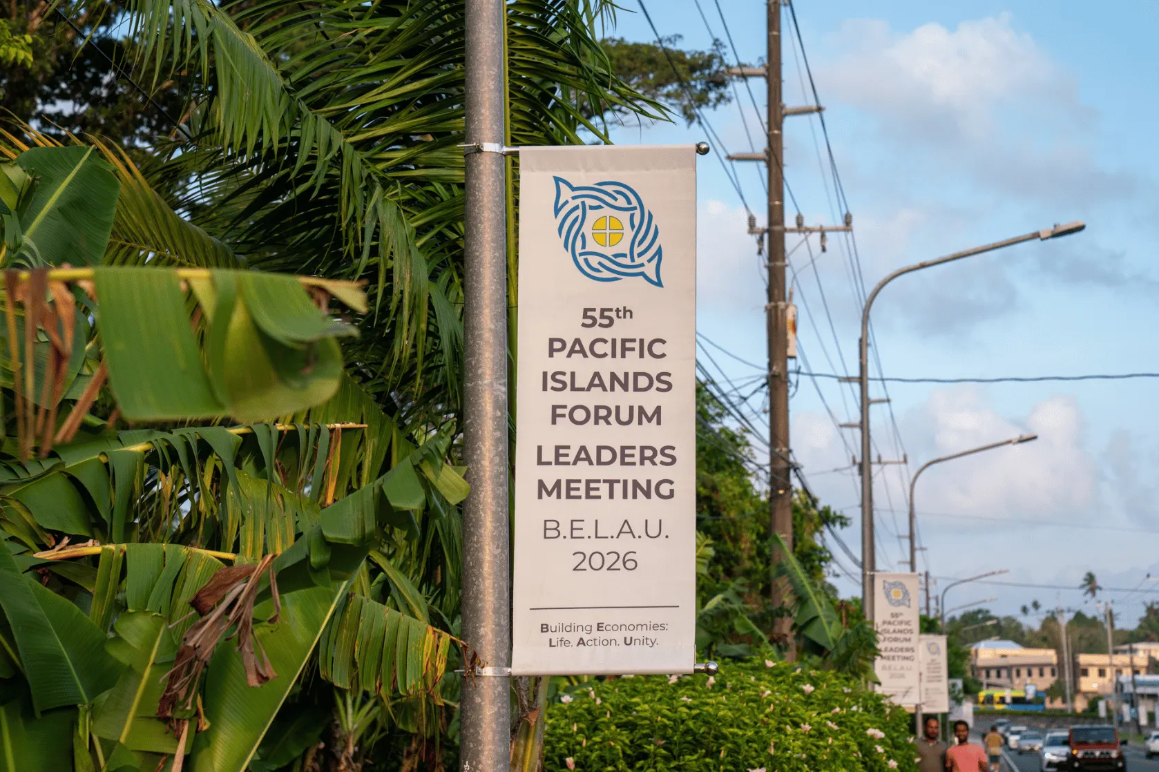 A banner for the 55th Pacific Islands Forum Leaders Meeting in Belau, 2026, hangs from a streetlight pole along a tropical roadside lined with palm and banana trees, utility poles and buildings.