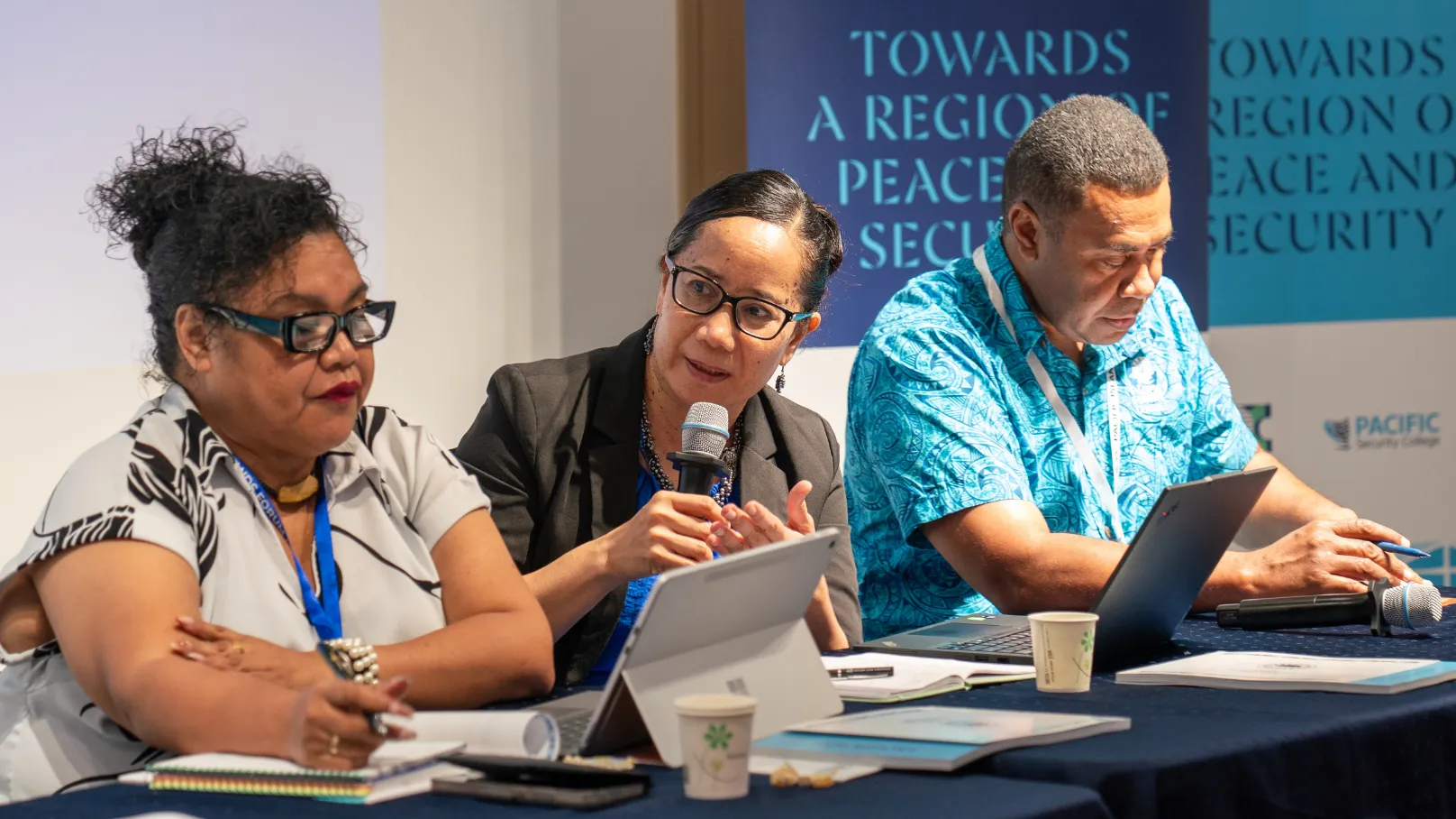 Three panel participants seated at a conference table during a regional peace and security event. One participant speaks into a handheld microphone while others take notes and use laptops and tablets.