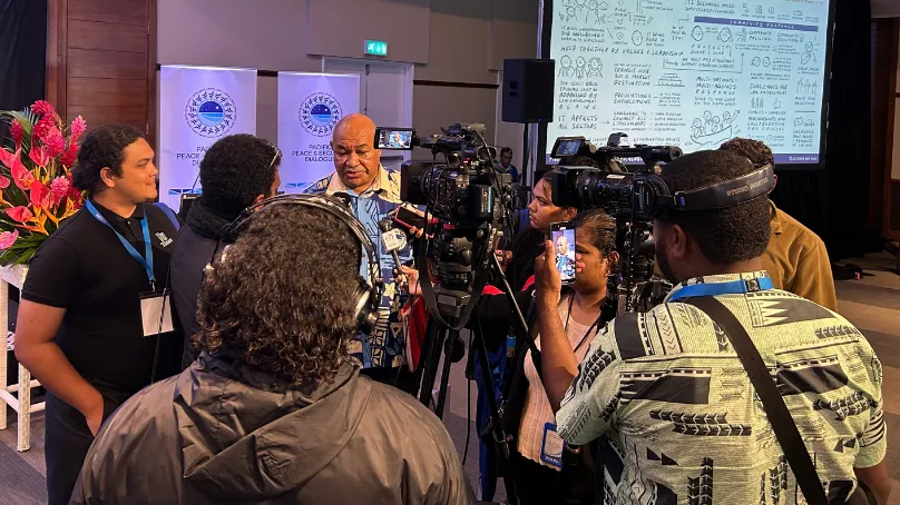 Media gather around a speaker who is wearing a Pacific Bula shirt during an interview at a Pacific peace and security event, with several television cameras and journalists recording. A presentation screen in the background displays illustrated notes on community cooperation and addressing transnational organised crime.