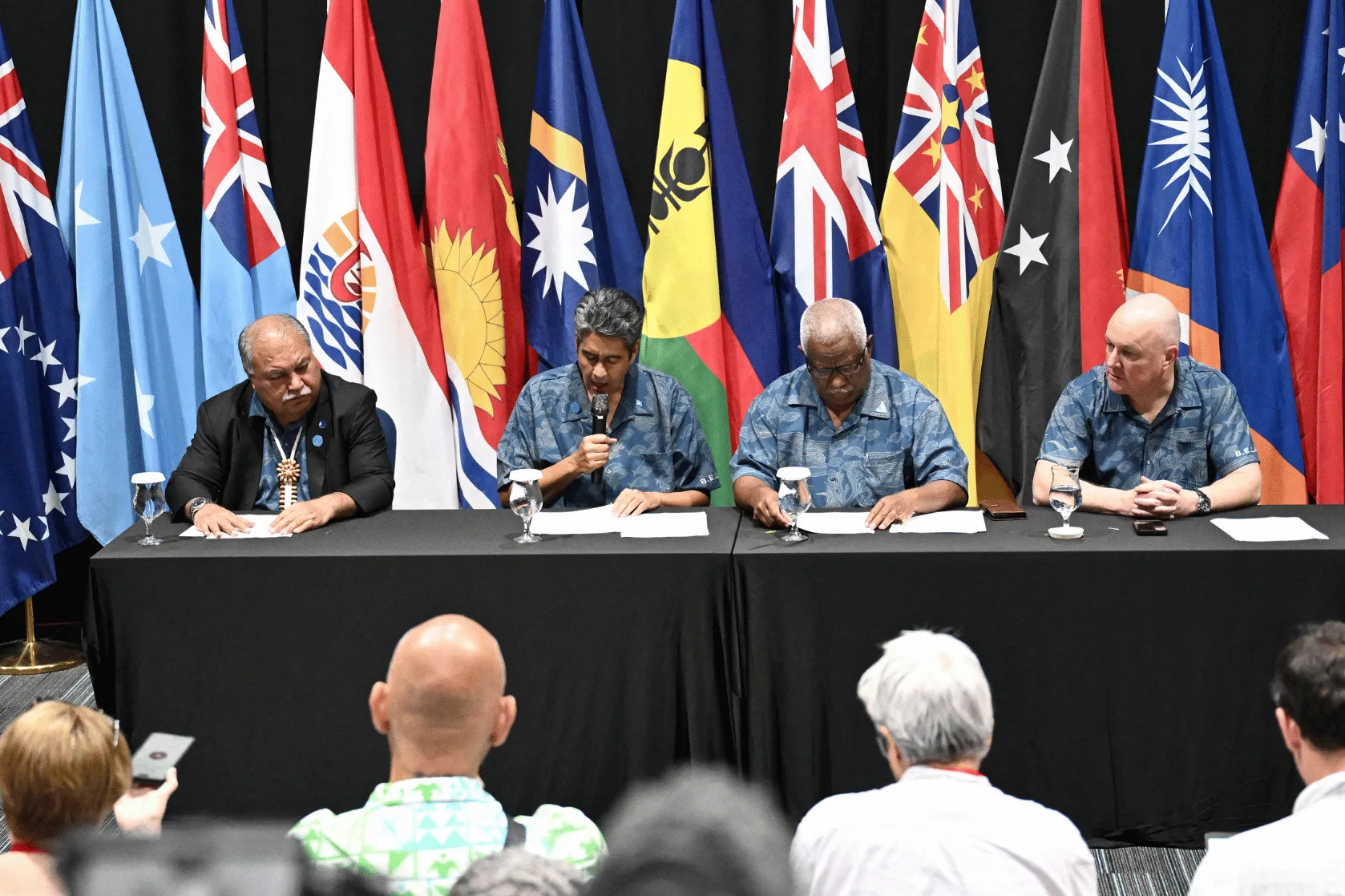Four people sit on a panel at a Pacific regional meeting, with flags from Pacific Island countries and territories displayed behind them. One speaker holds a microphone while the others listen.
