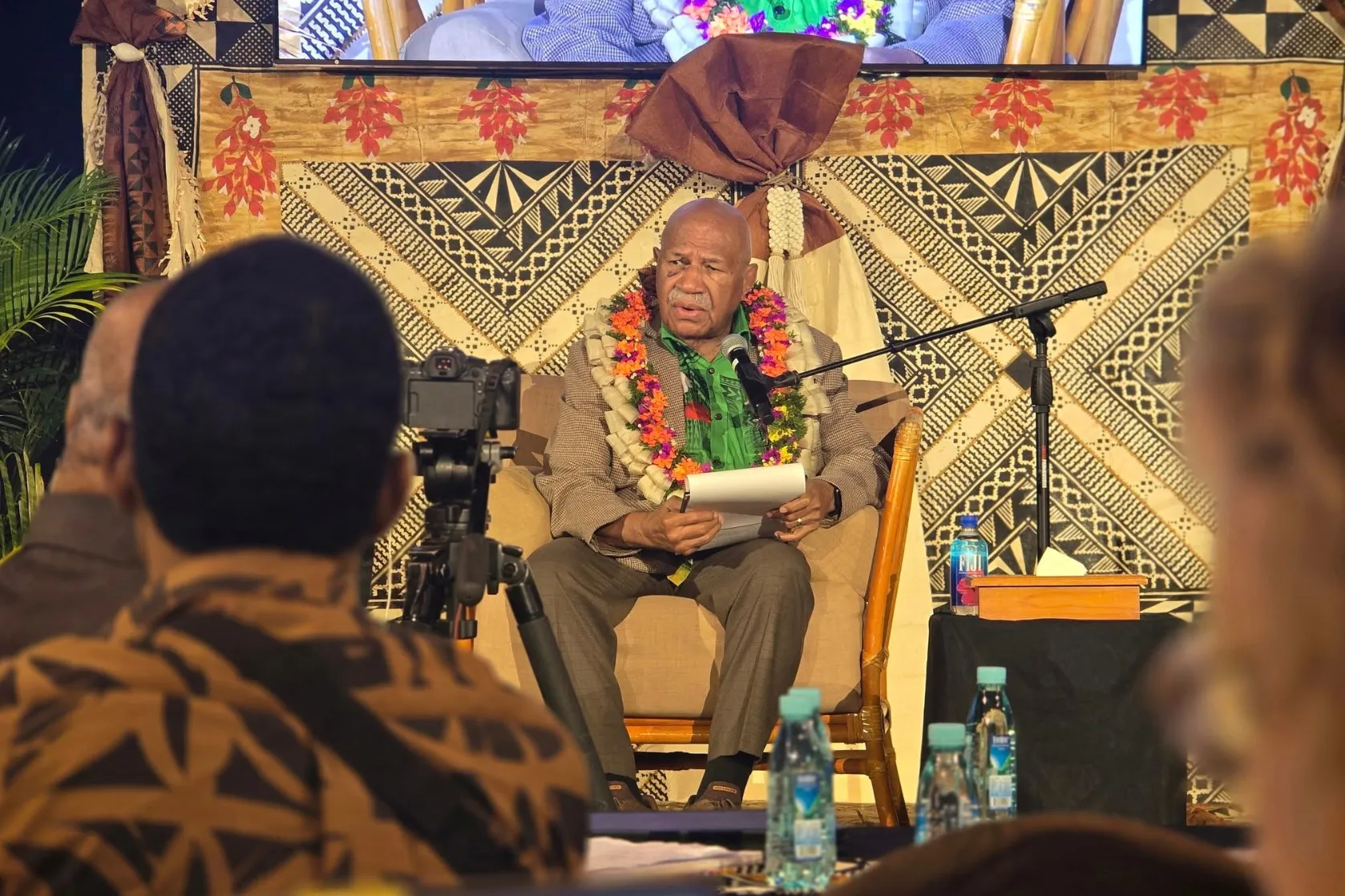 Fiji Prime Minister Sitiveni Rabuka speaks into a microphone while seated on stage at the Pacific Media Summit, surrounded by traditional Pacific decorations and floral garlands. Audience members and a video camera are visible in the foreground.