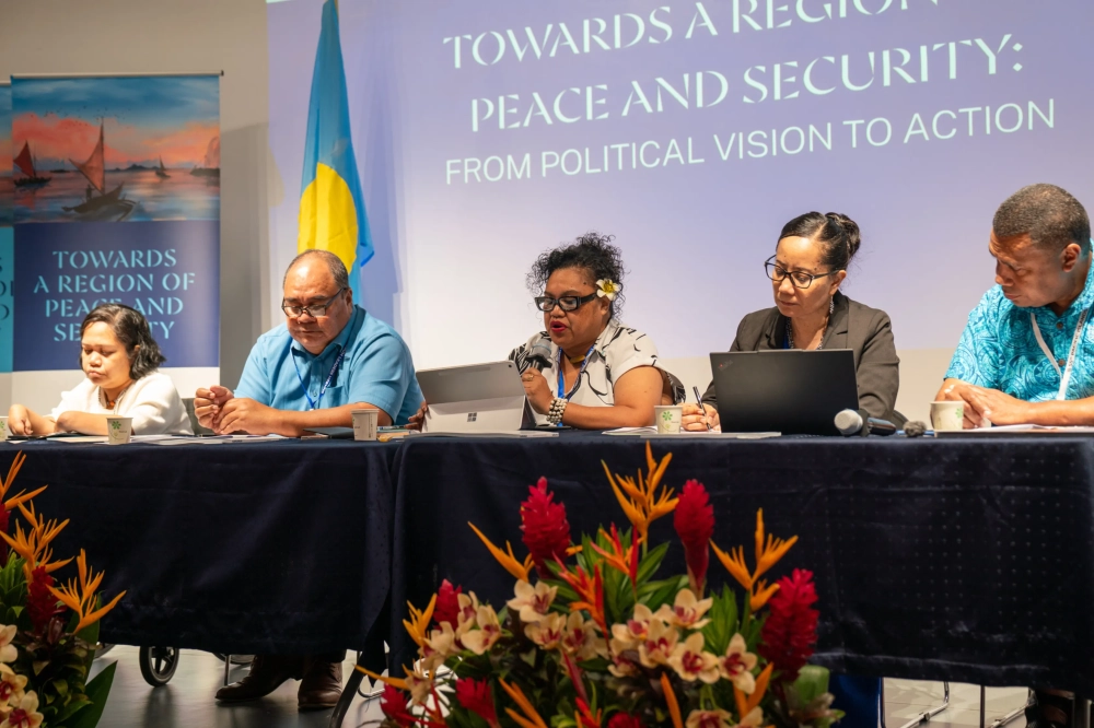 Panel discussion at a regional peace and security conference, showing five participants seated at a table with laptops, documents, and microphones. One participant addresses the audience while speaking into a microphone.