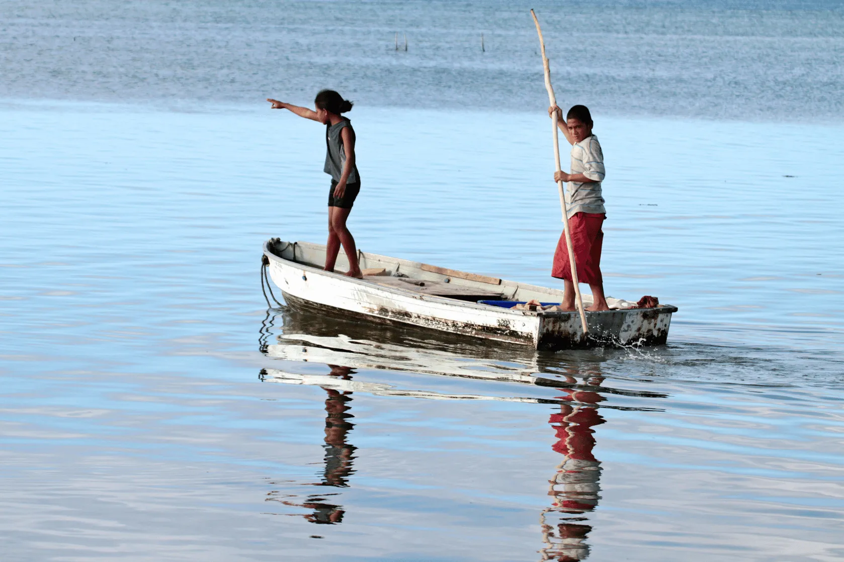 Two children stand in a small, weathered boat on calm blue water. One points towards the distance while the other holds a long wooden pole. Their reflections ripple across the water.