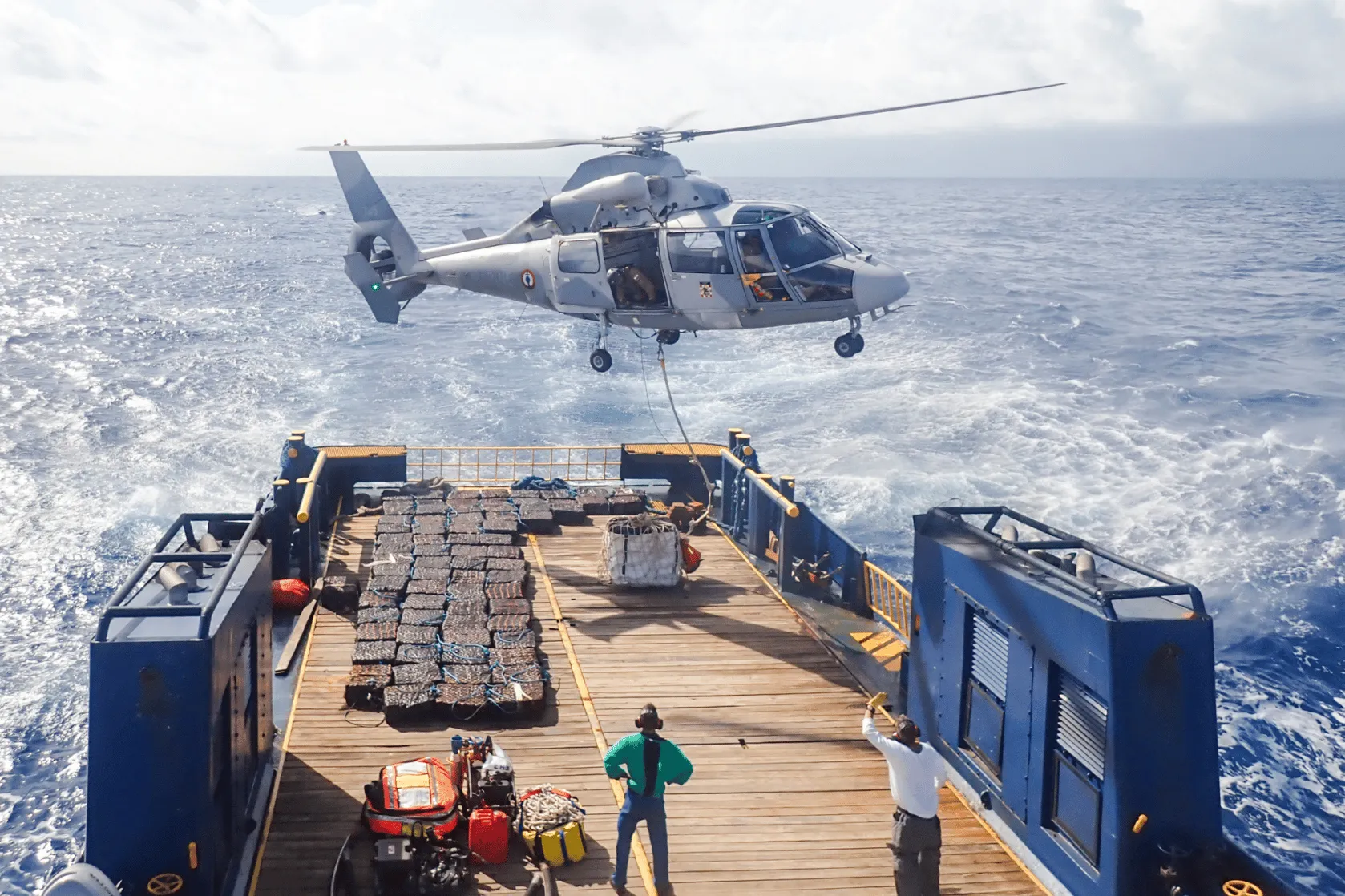 helicopter hovers above a vessel at sea as a cable is lowered from the aircraft to the vessel’s deck, where equipment and bundled materials are secured.