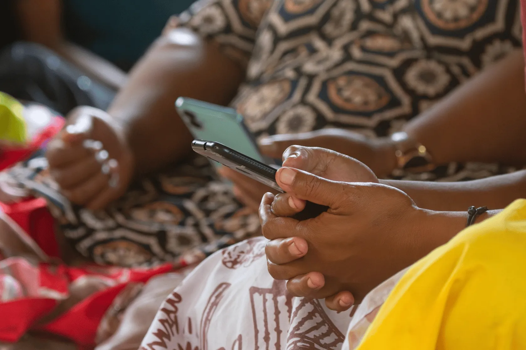 Close-up of a person holding a smartphone, with other people seated nearby in colourful clothing, including patterned Pacific-style fabrics.