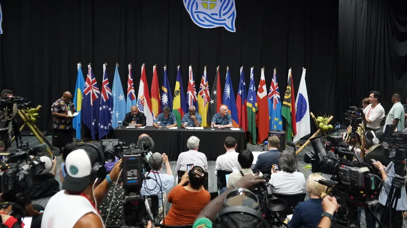 Pacific Islands Forum leaders and representatives sit at a conference table in front of national flags during a media conference, as journalists and camera operators record the event.