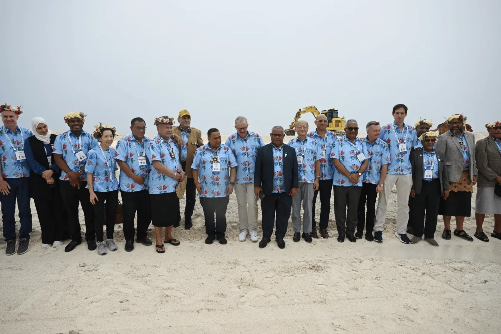 A group of Pacific and international delegates pose for a photograph at an outdoor construction site, wearing matching light blue floral shirts, traditional floral head garlands and formal attire. A yellow excavator is visible in the background.