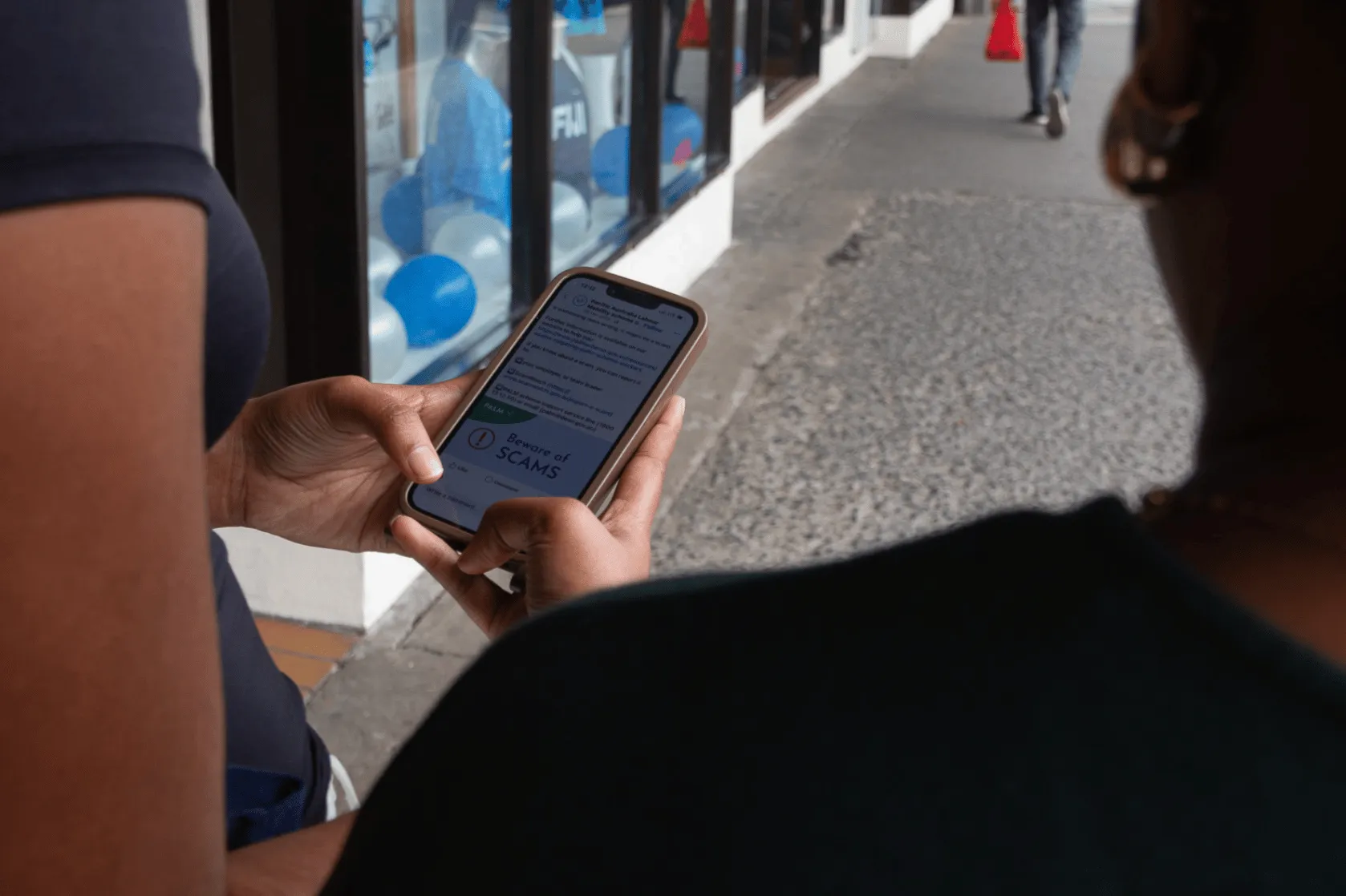 A person uses a smartphone to view a social media post warning about scams while standing on a footpath outside a shop.